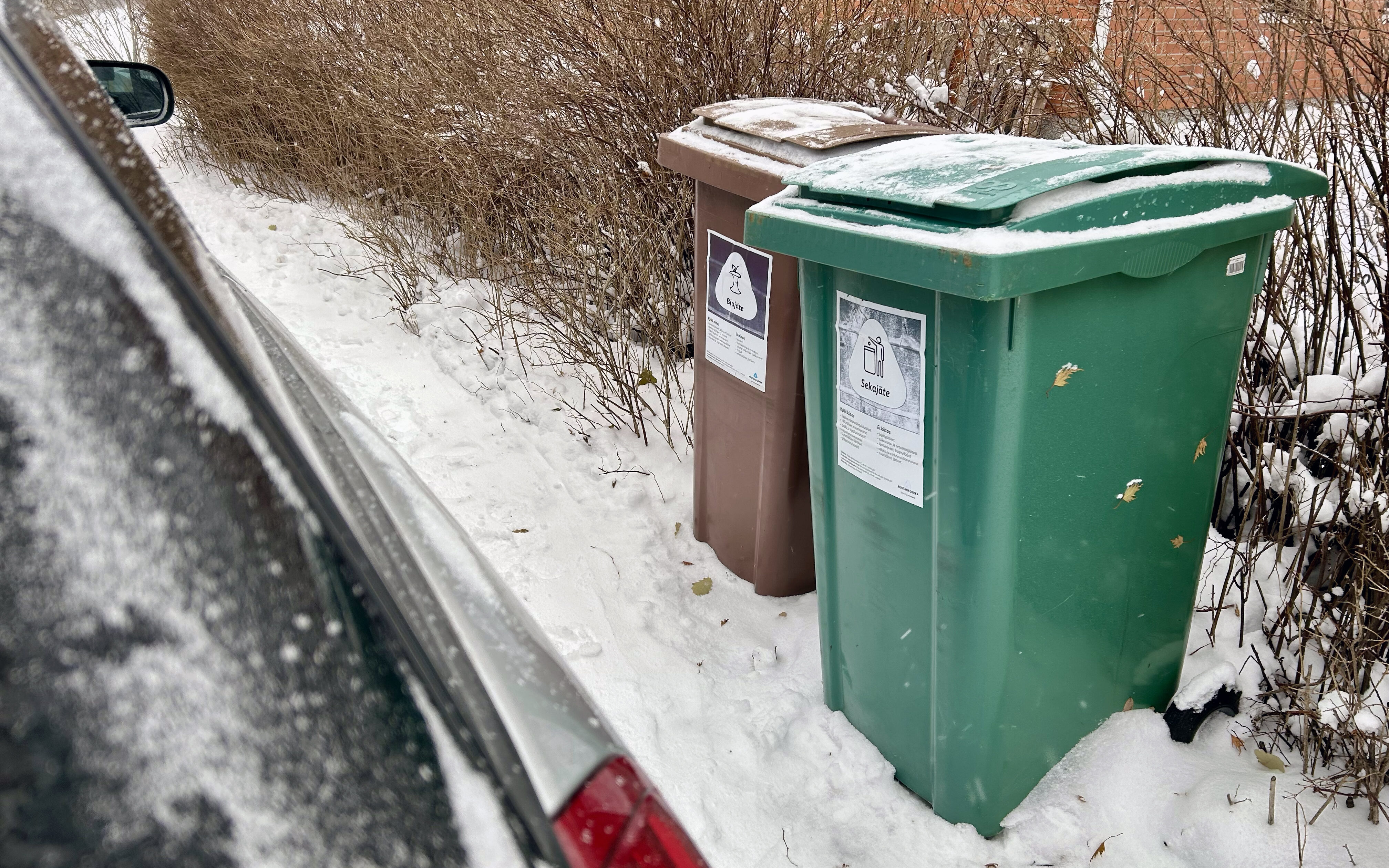 Vasemmalla näkyy osa harmaasta autosta ja oikealla kaksi jäteastiaa, sekajätteelle ja biojätteelle. Auton ja astian välissä on melko pieni tila.