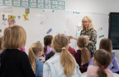 Vaaleahiuksinen opettaja kertoo luokalle jotain. Lapset ovat kameraan selin.