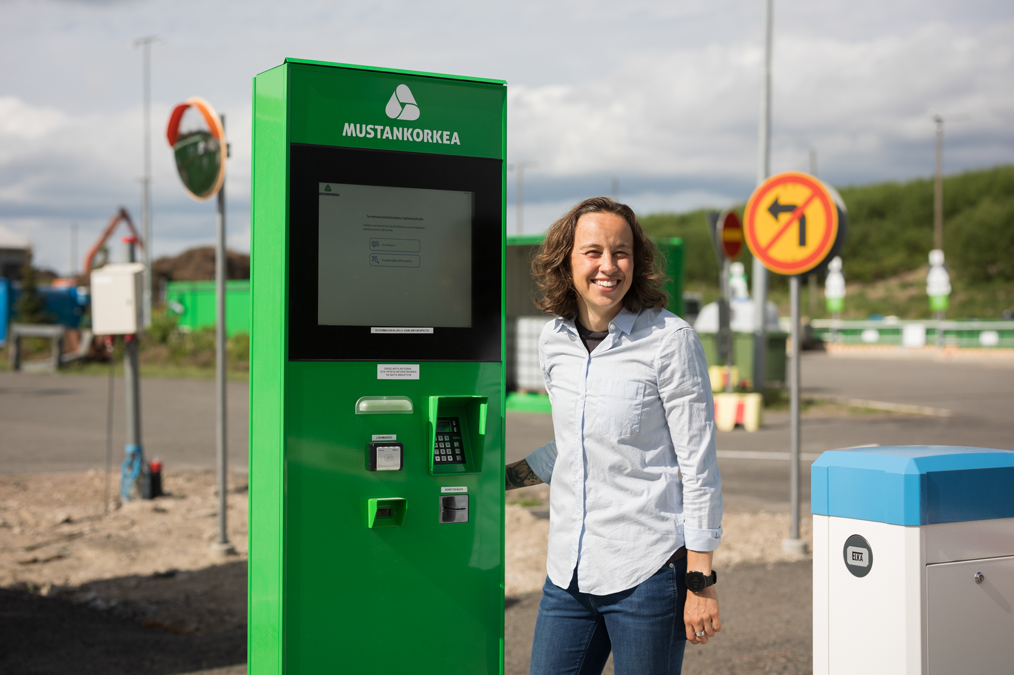 Nainen seisoo vihreän automaatin vieressä. Takana näkyy liikennemerkkejä ja lajittelupihaa.