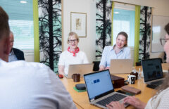 Kaksi naistaa istuu vierekkäin ja nauraa. Etualalla näkyy sivusta naisen kasvoja ja takaa miehen selkää. Ihmiset ovat pitämässä palaveria neuvotteluhuoneessa.