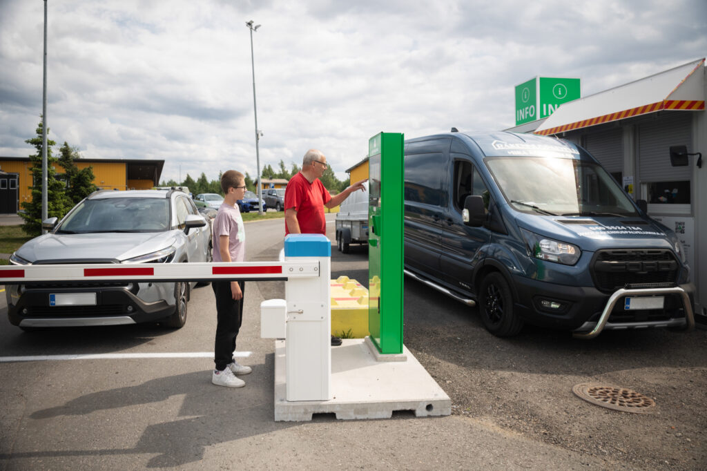 Kaksi autoa on rinnakkain. Punapaitainen vanhempi mies asioi vihreällä automaatilla. Hänen takanaan seisoo nuorukainen. Oikealla on pakettiauto peräkärryn kanssa infopisteellä.