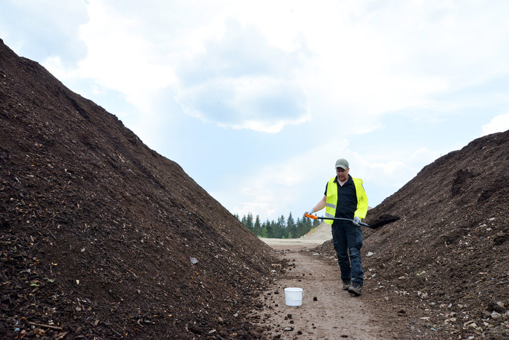 Huomioliivit päällä oleva mies kantaa multaa lapiossa valkoiseen sankoon. Taivaalla näkyy hieman pilviä ja miehen vieressä on kaksi kompostiaumaa.