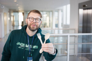 Vihreäpaitainen mies pitää käsissään puista kertakäyttöhaarukkaa ja -lusikkaa. Taustalla näkyy yliopiston yläaulaa.