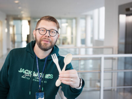 Vihreäpaitainen mies pitää käsissään puista kertakäyttöhaarukkaa ja -lusikkaa. Taustalla näkyy yliopiston yläaulaa.