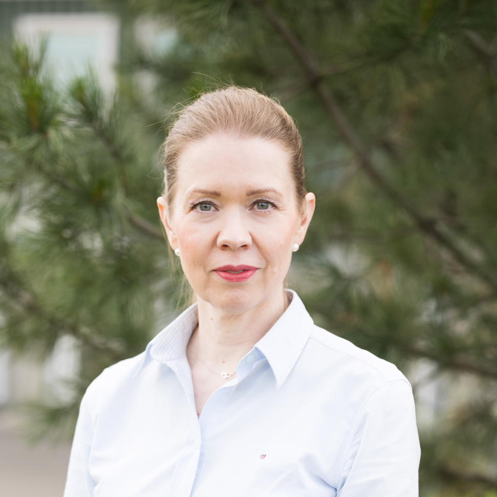 Nainen kauluspaidassa katsoo kameraan ja hymyilee vienosti. Takana näkyy vihreää puuta.