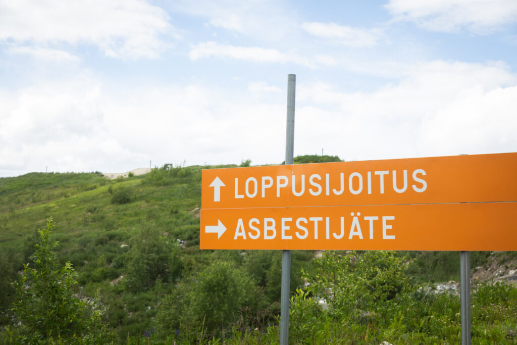 Oransseissa kylteissä lukee loppusijoitus ja asbesti. Takana näkyy vihreää nurmikkoa.