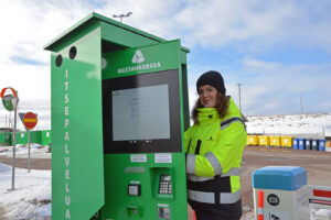 Nainen on itsepalveluautomaatin vieressä. Automaatin ovi on kiinni ja naisen kädet ovat jossain sen takana. Etualalla näkyy maksupääte.