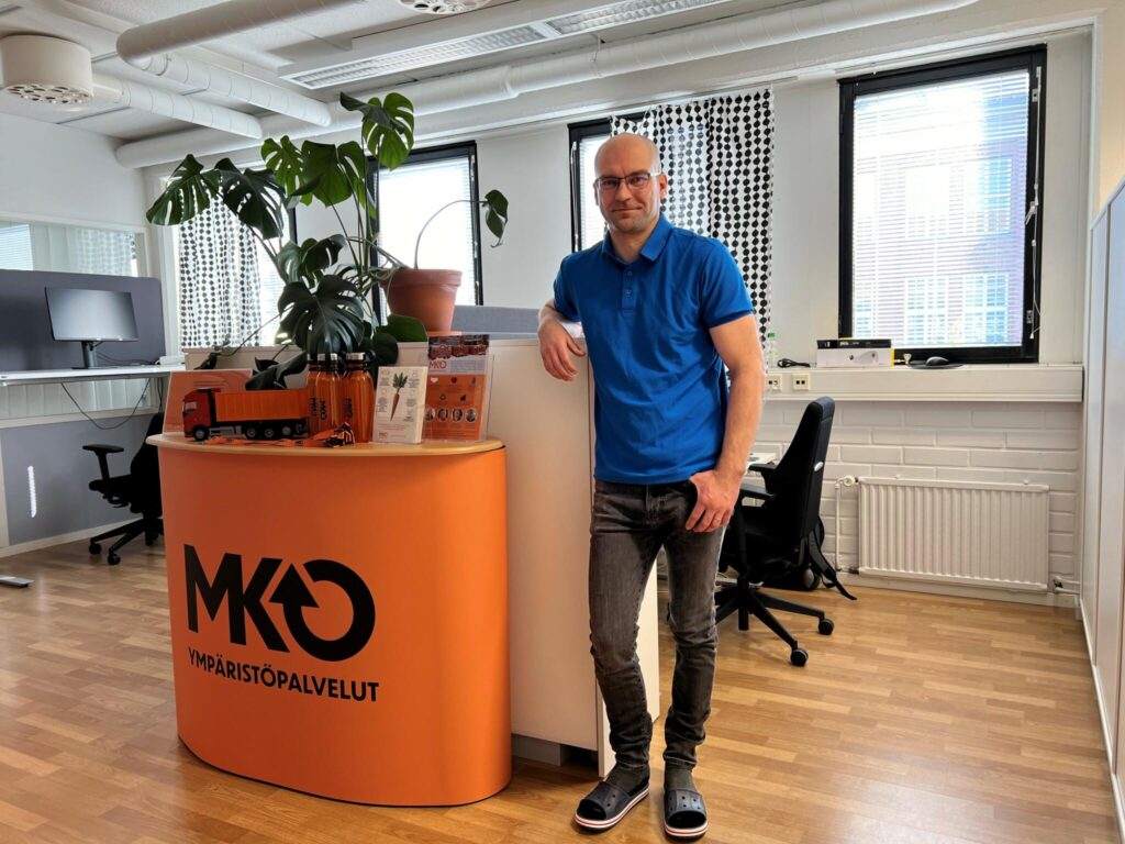 Mies seisoo nojaten pöytään ja hymyilee. Hän on toimistossa, jossa näkyy oranssi ständi ja toimistokalusteita.