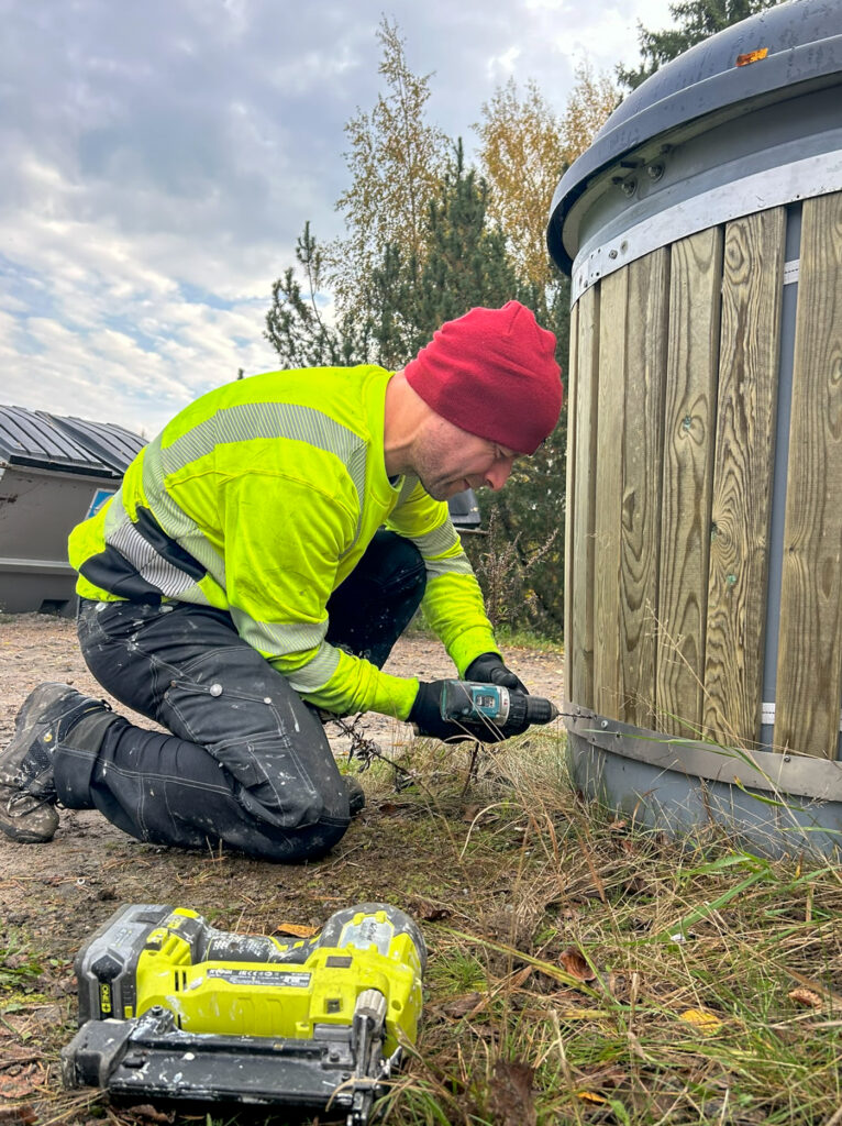 Mies poraa hyötykeräyspisteen pohjalistaa kiinni. Miehellä on keltainen huomiopaita ja punainen pipo.