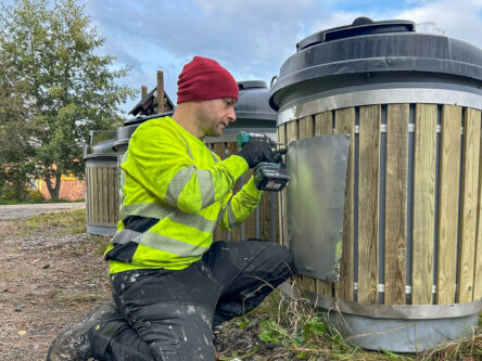 Mies poraa hyötykeräyspisteellä metallilaattaa pisteeseen kiinni. Miehellä on keltainen huomiopaita ja punainen pipo.