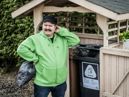 Mies raapii päätään tuskaisena. Takana näkyy jätekatos, jossa on sekajäteastia.