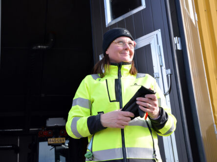 Nainen pitää kädessään mustia hanskoja ja katsoo eteenpäin hymyillen. Hänellä on päällään huomiovaatteet. Takan näkyy jokin varastorakennus ovi auki.