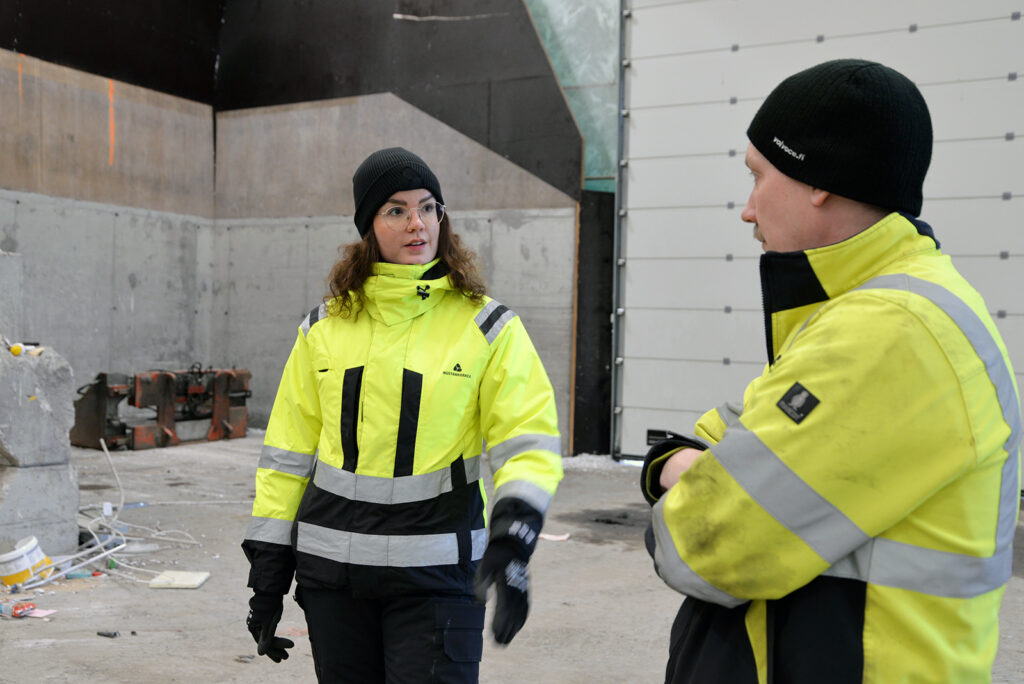 Nainen kertoo miehelle jotain ja osoittaa hieman kädellä. Kummallakin on päällään huomiovaatteet. He ovat jossain hallitilassa.