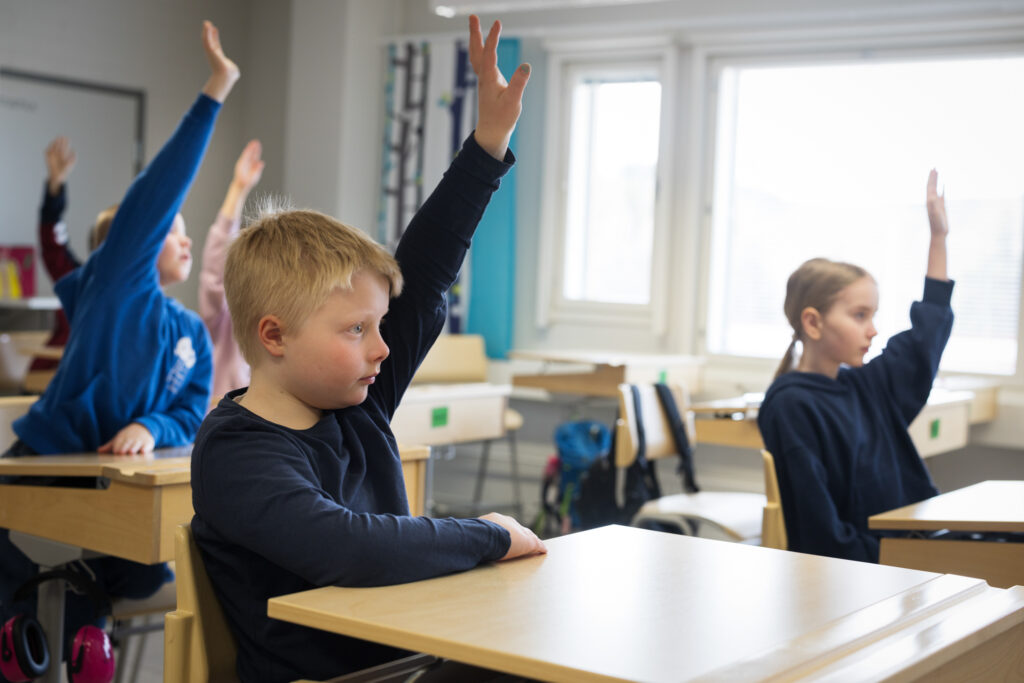 Viisi lasta viittaa koululuokassa. Etualalla on poika, hänen takanaan näkyy sumeasti tyttö. Luokassa on myös muita lapsia.