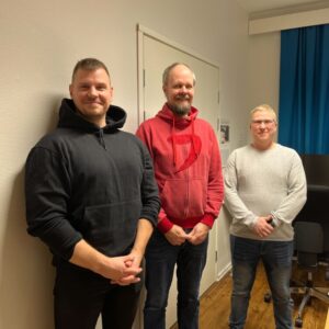 Kolme miestä seisoo rinnakkain jossain toimistohuoneessa, jonka pöydältä pilkottaa pieni joulukuusi. Miehiä naurattaa.