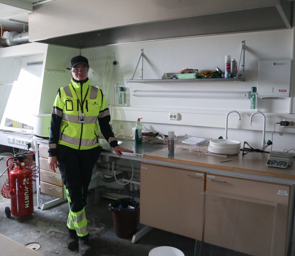 Henkilökuva Salla Leskisestä. Hänellä on päällä keltainen huomiotakki, lippalakki sekä suojalasit. Hän nojaa pöytään ja katsoo kameraan.