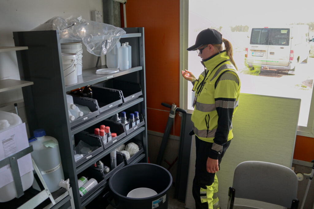 Nainen seisoo ja katsoo tarvikehyllyä. Hänellä on päällä keltainen huomiotakki, lippalakki sekä suojalasit.
