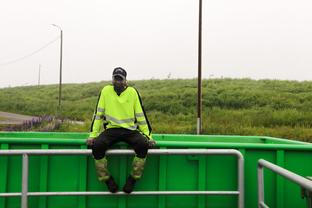 Mies istuu vihreän jätelavan reunalla. Hän hymyilee ja katsoo kameraan. Hänellä on päällään työvaatteet ja musta lippis.