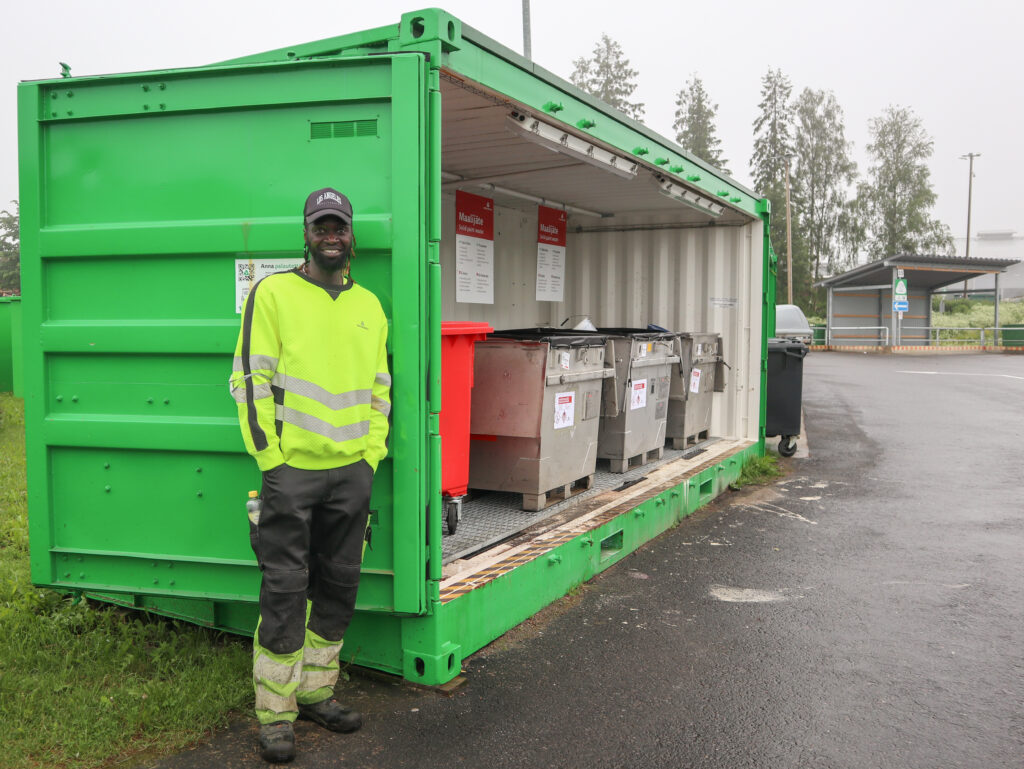Mies seisoo vaarallisen jätteen kontin edessä hymyillen kädet taskuissa. Hänellä on päällään keltainen työpaita ja musta lippis. Taustalla näkyy maalijätteen jäteastioita ja lajittelupiha.