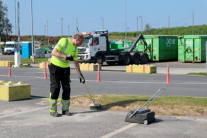 Mies huomiovaatteissa lakaisee lajittelupihalla. Vieressä on keltaisia betoniporsaita. Takana näkyy jätekeskuksen aluetta.