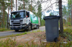Etualalla näkyy harmaa jäteastia puun alla. Taustalla näkyy Mustankorkean jäteauto.