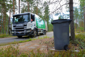 Etualalla näkyy harmaa jäteastia puun alla. Taustalla näkyy Mustankorkean jäteauto.