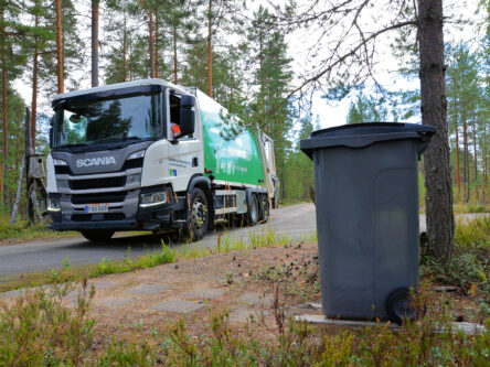 Etualalla näkyy harmaa jäteastia puun alla. Taustalla näkyy Mustankorkean jäteauto.