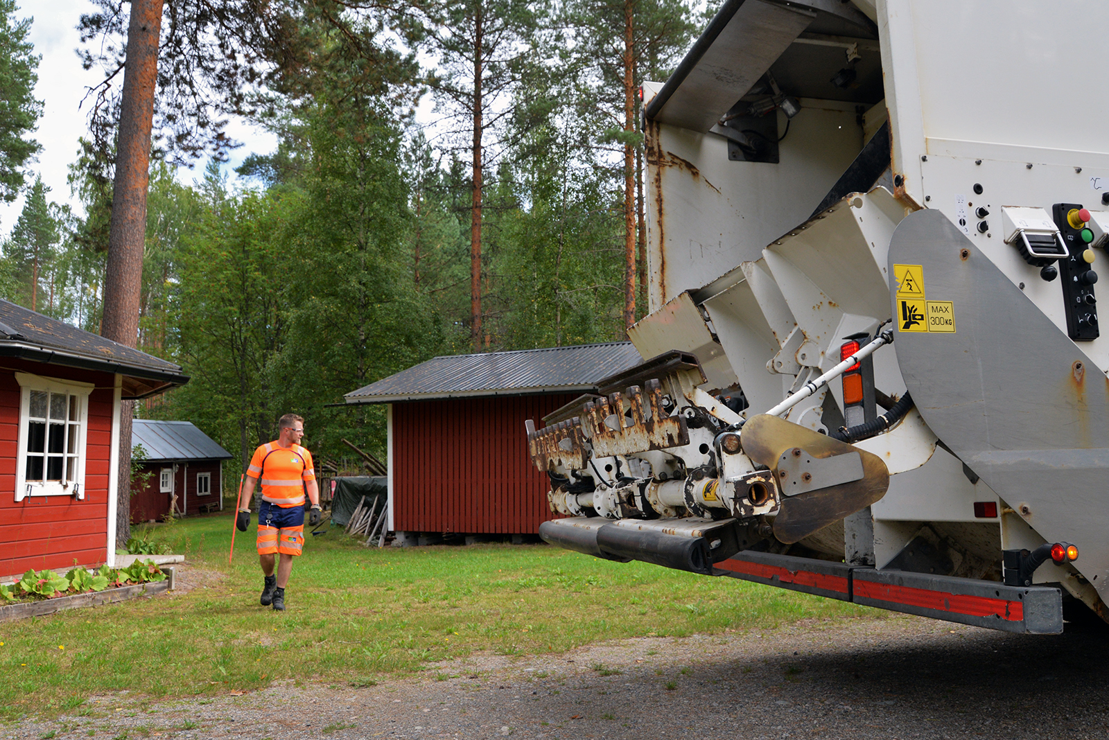 Huomiovaatteet päällä oleva mies tähyilee jonkin mökkipihan ympäristöä. Jäteauton perää näkyy oikealla.