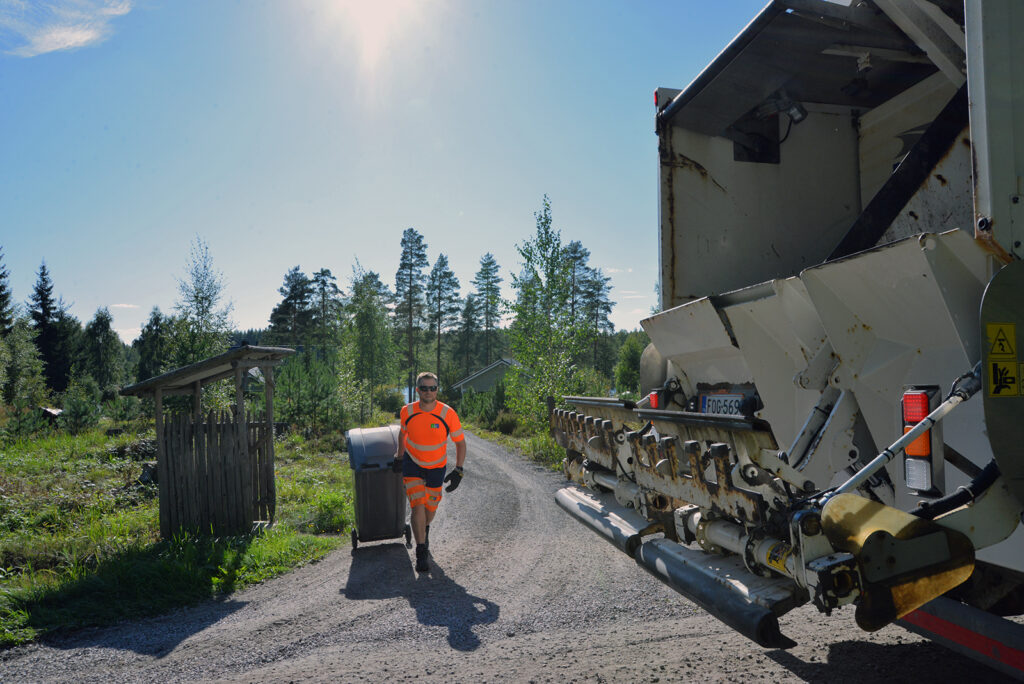 Huomiovaatteet päällä oleva mies vetää jäteastiaa jäteautolle. Takana näkyy hieman taloa ja järveä. On kaunis kesäpäivä.