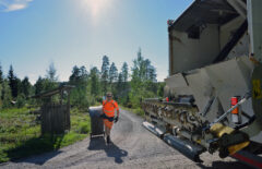 Huomiovaatteet päällä oleva mies vetää jäteastiaa jäteautolle. Takana näkyy hieman taloa ja järveä. On kaunis kesäpäivä.