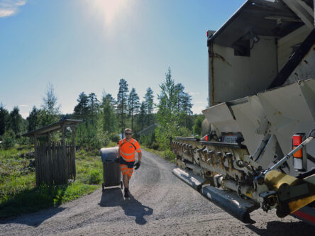 Huomiovaatteet päällä oleva mies vetää jäteastiaa jäteautolle. Takana näkyy hieman taloa ja järveä. On kaunis kesäpäivä.