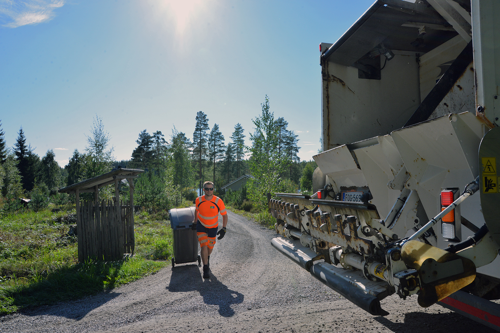 Huomiovaatteet päällä oleva mies vetää jäteastiaa jäteautolle. Takana näkyy hieman taloa ja järveä. On kaunis kesäpäivä.
