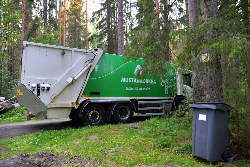 Etualalla näkyy harmaa jäteastia ja sen takana Mustankorkean jäteauto. On kaunis kesäinen päivä jossain metsikössä.
