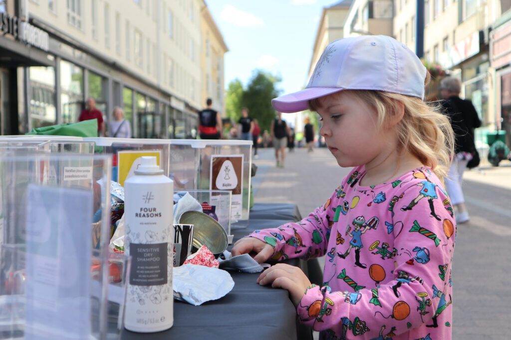 Lapsi lajittelee mustankorkean pisteellä Kävelykatukirppiksellä. Lapsella on päällä pinkki paita, jossa on kuvia Peppi Pitkätossusta. Edessä on erilaisia jätteitä, joita voi lajitella pieniin jäteastioihin.