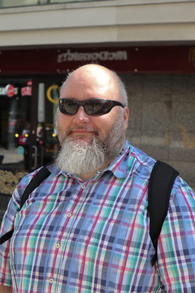 Mies katsoo kameraan mustat aurinkolasit päässä. Hänellä on pitkä harmaa parta ja sininen ruutupaita päällä.
