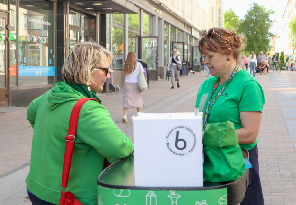 Kaksi naista keskustelevat Mustankorkean pisteellä Kävelykatukirppiksellä. Molemmilla on vihreät paidat päällä. Taustalla näkyy Jyväskylän Kauppakatu.