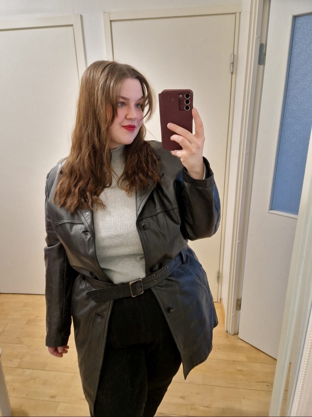 Nainen poseeraa kuvaan peilin kautta. Hänellä on päällään musta nahkatakki ja hän ottaa kuvaa itse puhelimellaan.
