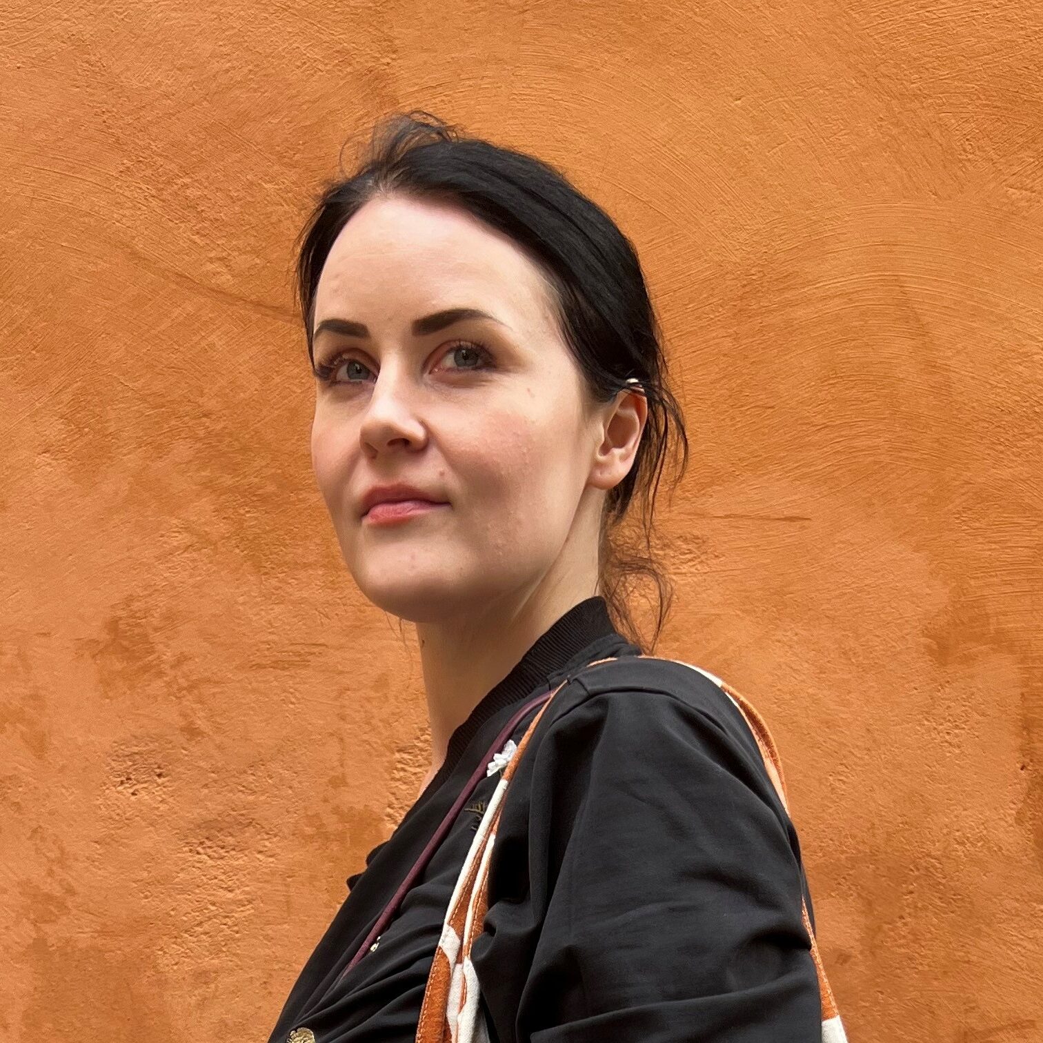 Tummatukkainen nainen seisoo oranssin seinän edessä ja katsoo kameraan. Hänellä on jalassaan raidalliset housut ja olalla oranssi marimekon unikkokuvioinen kangaskassi.