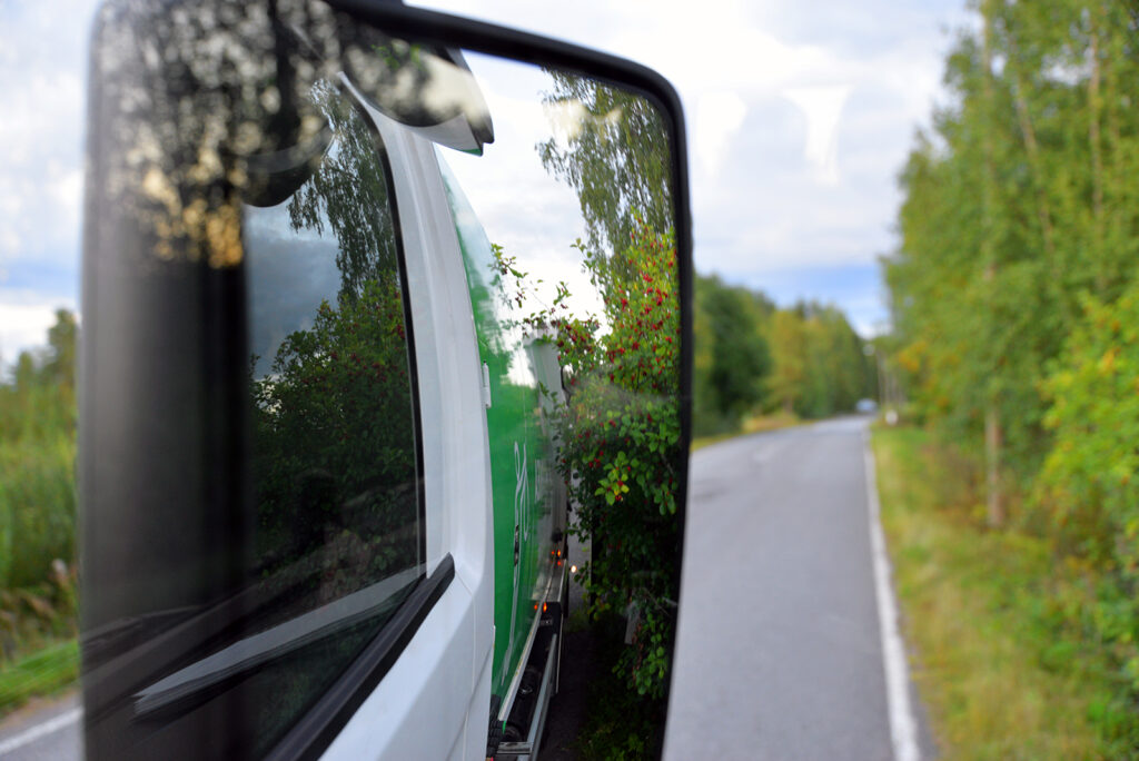 Jäteauton sivupeilistä näkyuy oksia, jotka raapivat jäteuatoa. Taustalla näkyy asfalttitietä.
