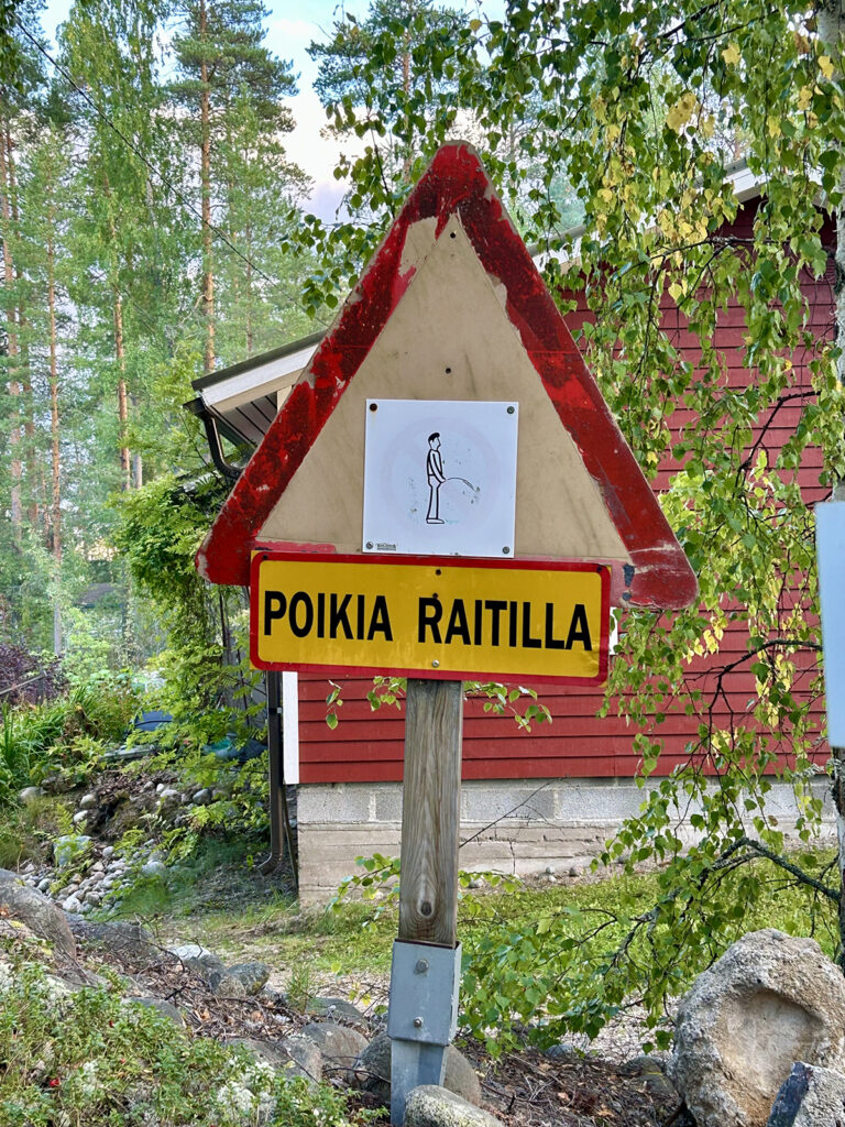 Puinen kolmiomallinen liikennemerkki, jossa on piirroskuva miehestä pissaamassa ja alla lukee "poikia raitilla".