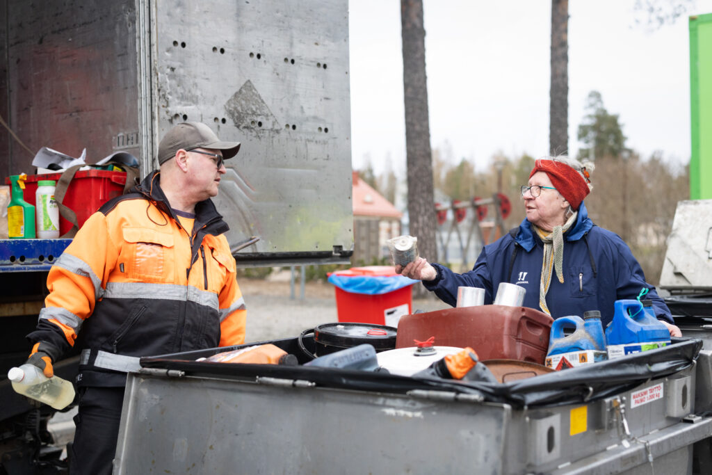 Nainen näyttää miehelle jotain pientä metallipönttöä kysyvästi. Edessä on iso keräysastia erilaisia purkkeja, joissa joissain näkyy vaarallisten jätteiden huomiomerkki.