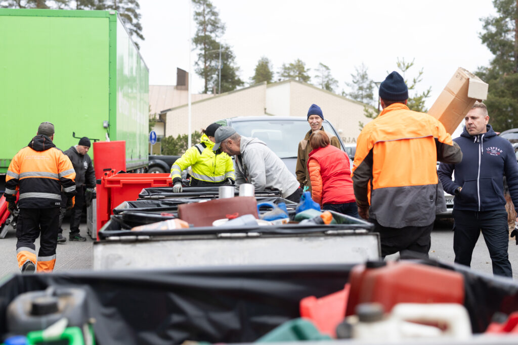 Keskellä näkyy erilaisia isoja metallia keräyslaatikoita ja niiden ympärillä ihmisiä. Joillain on huomiovaatteet, mutta suuremmalla osalla ei. Eräs mies hymyilee. Kaikki ovat touhuissaan.