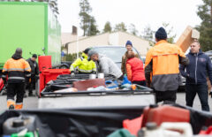 Keskellä näkyy erilaisia isoja metallia keräyslaatikoita ja niiden ympärillä ihmisiä. Joillain on huomiovaatteet, mutta suuremmalla osalla ei. Eräs mies hymyilee. Kaikki ovat touhuissaan.