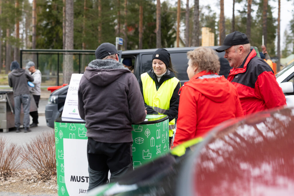 Huomioliivi päällä oleva nainen hymyilee. Hän seisoo ständillä, jonka luona on kolme asukasta.