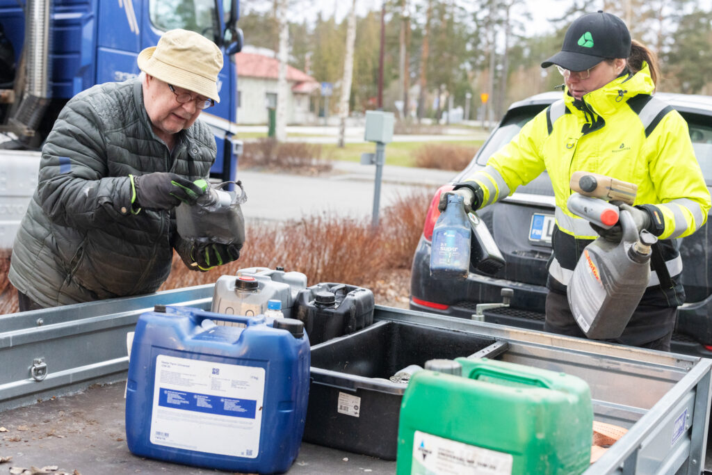 Huomiotakkinen nainen pitelee käsissään erilaisia kanistereita. Vieressä vanhempi mies tiirailee pullon merkintöjä. Etualalla näkyy eirlaisia öljykanistereita.