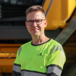 Mies seisoo pyöräkuromaajan edessä ja katsoo kameraan. Hänellä on mustasankaiset silmälasit ja mustakeltaiset työvaatteet päällä.