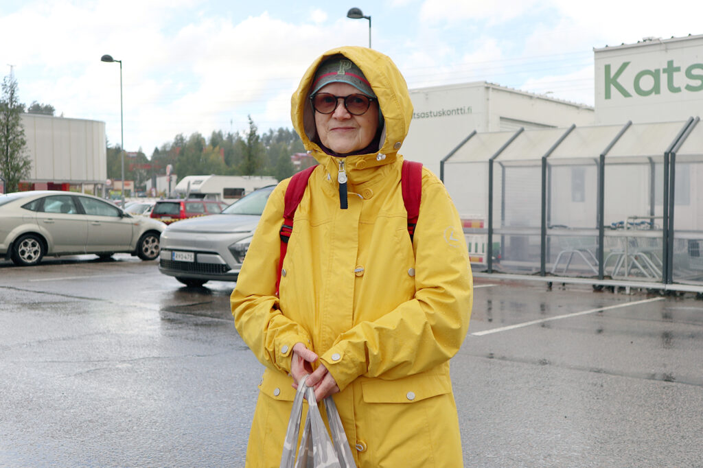 Keltainen takki päällä oleva nainen katsoo kameraan huppu päässään. Hänellä on käsissään jokin muovipussi.