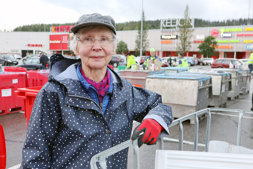 Nainen pilkullisessa sadetakissa nojailee jäteastiaan. Takana näkyy isoa kauppakeskusta.