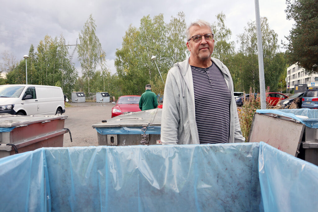Raitapaitainen mies seisoo jonkin jäteastian takana. Taustalla näkyy puita ja henkilöautoja.