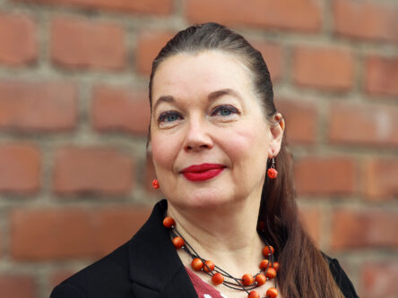 Nainen hymyilee ja katsoo kameraan. Takana näkyy punatiiliseinää. Naisella on päällä musta bleiseri ja punavalkoinen paita.